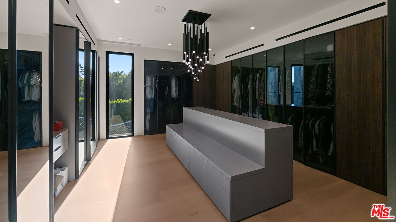 This is a spacious walk-in closet featuring custom cabinetry with glass doors and wood paneling. A central island with drawers and shelving provides additional storage and display space. A modern chandelier hangs from the ceiling, illuminating the organized clothing and accessories, creating a luxurious and functional space.