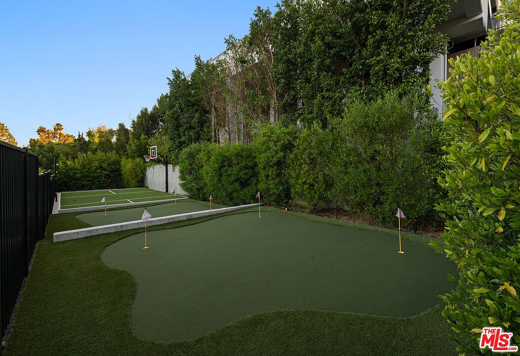 This image showcases a meticulously maintained yard featuring a putting green and a basketball court. The lush green turf and surrounding mature trees create a private and serene outdoor space. The area is well-defined with concrete borders and fencing, suggesting a high-end property with recreational amenities.