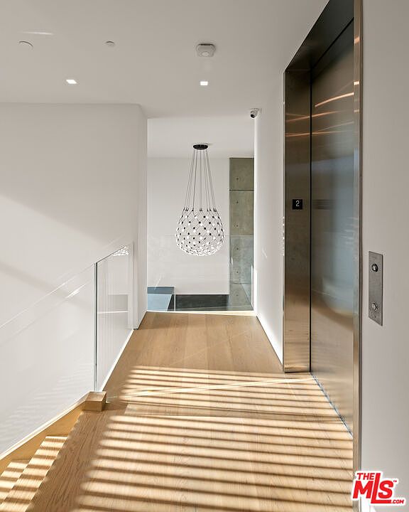 This interior shot showcases a modern hallway with light wood flooring and white walls. A striking pendant light fixture hangs in the center, while a stainless steel elevator door is visible on the right. The hallway leads to a bright space, creating a clean and contemporary aesthetic.