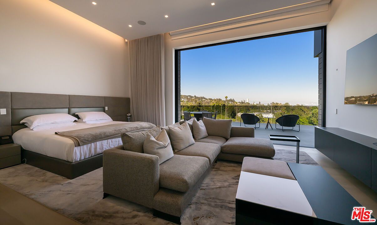 This is a primary bedroom featuring a large bed with a gray headboard, a gray sectional sofa, and a modern coffee table. A large window offers a view of the outdoors, and a sleek entertainment center is mounted on the wall. The room has a luxurious and contemporary feel.