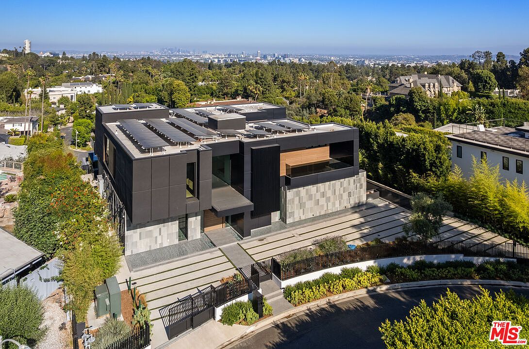 This aerial view showcases a modern, multi-level home with a striking dark exterior and solar panels on the roof. The property features a meticulously designed driveway with linear concrete slabs and lush landscaping. The house is nestled among mature trees, offering privacy and a sense of seclusion, with a cityscape visible in the distance.