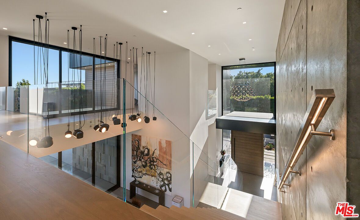 This interior shot showcases a modern hallway and staircase with a focus on architectural details. The design features a glass railing, a striking concrete wall with integrated lighting, and a unique cluster of pendant lights hanging above the stairs. Large windows provide ample natural light, highlighting the clean lines and contemporary aesthetic of the space.
