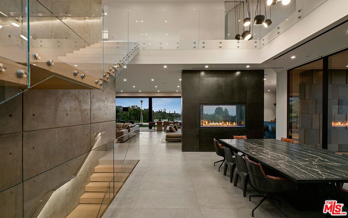 This interior shot showcases a modern dining area with a long, dark marble table and sleek black chairs. A striking concrete staircase with glass railings is visible on the left, while a contemporary fireplace adds warmth and ambiance to the right. Large windows in the background offer a glimpse of the outdoor scenery, enhancing the room's spacious and luxurious feel.