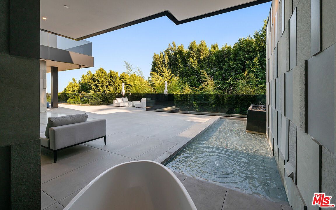 This image showcases a luxurious outdoor patio area, featuring sleek, modern design elements. A minimalist gray sofa sits on the expansive tiled patio, adjacent to a shallow water feature that runs along a textured wall. In the background, outdoor seating is visible beyond a glass barrier, all set against a backdrop of lush greenery, creating a serene and sophisticated outdoor living space.