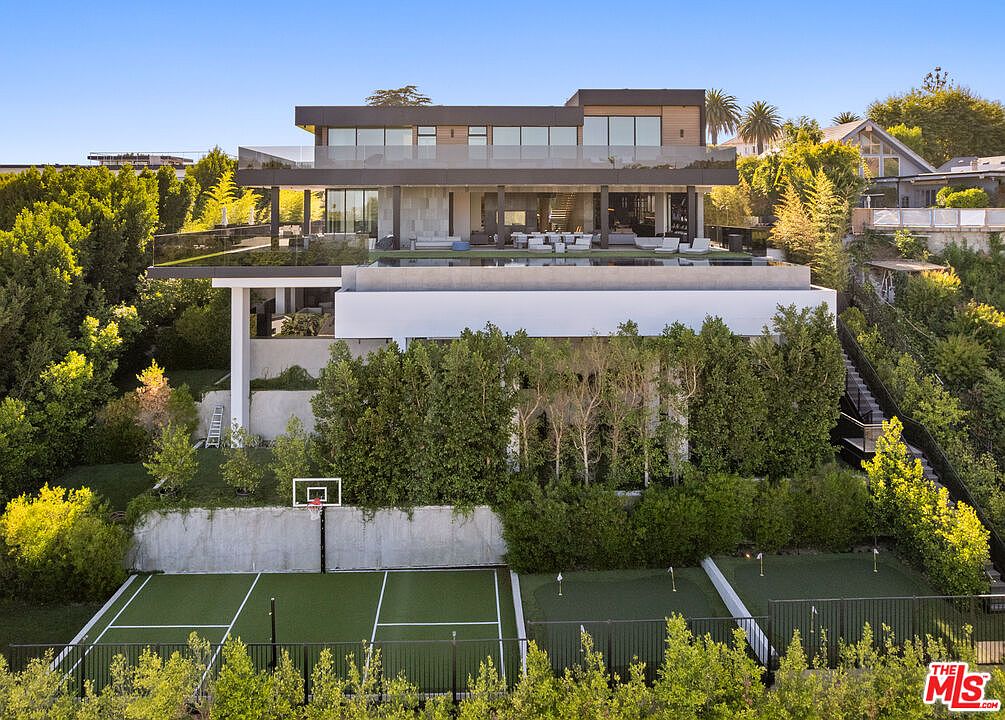 This image showcases the rear exterior of a modern luxury home. The multi-level structure features expansive glass windows and doors, a pool area with lounge seating, and a well-manicured yard with a tennis court and putting green. The property is surrounded by lush greenery, providing privacy and a serene atmosphere.