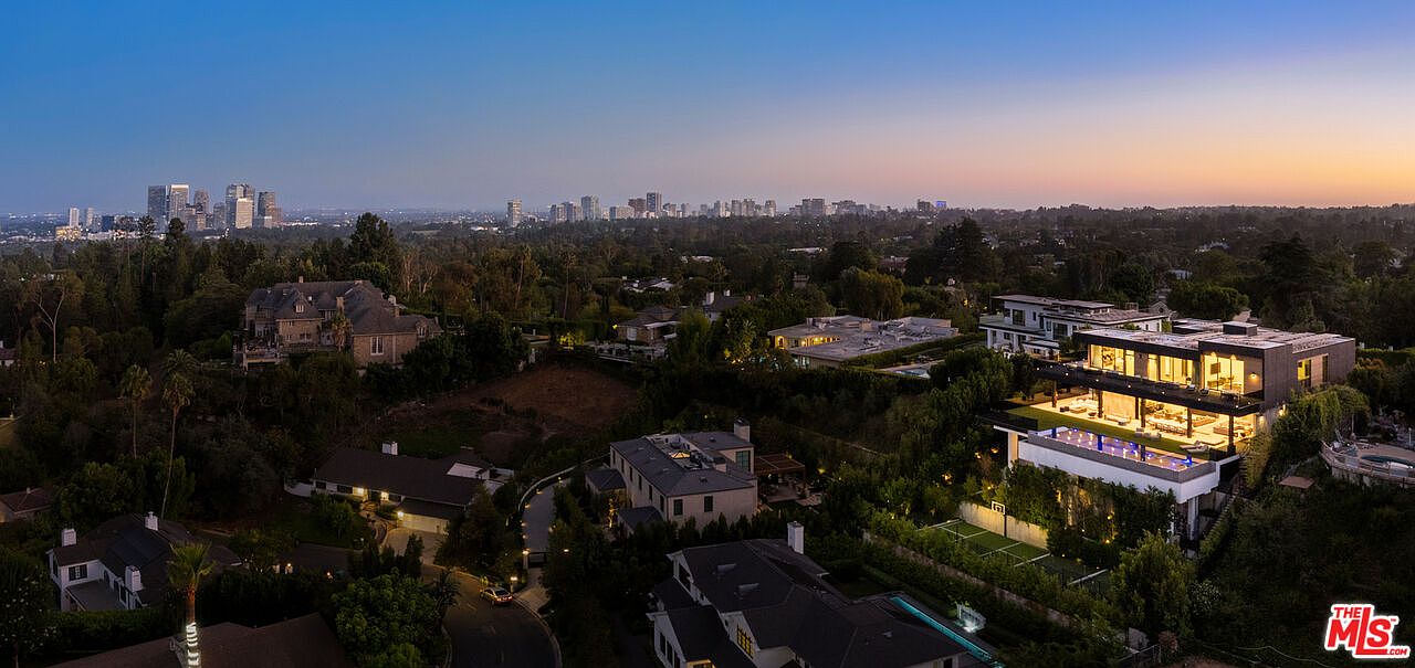 This aerial view showcases a luxurious residential area at dusk, featuring multiple modern homes with illuminated interiors and exteriors. The properties are surrounded by lush greenery, with a cityscape visible in the distance under a gradient sky. The image emphasizes the upscale nature and serene setting of the real estate.