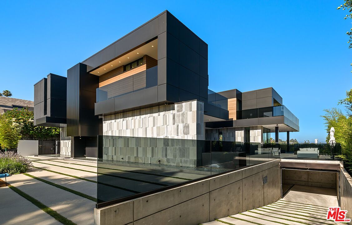 This is a striking front exterior view of a modern, multi-story home. The architectural design features a combination of dark, rectangular volumes and lighter, textured wall panels, creating a visually dynamic facade. A reflecting pool adds to the contemporary aesthetic, and the driveway is paved with concrete slabs interspersed with grass, leading to a hidden garage entrance.