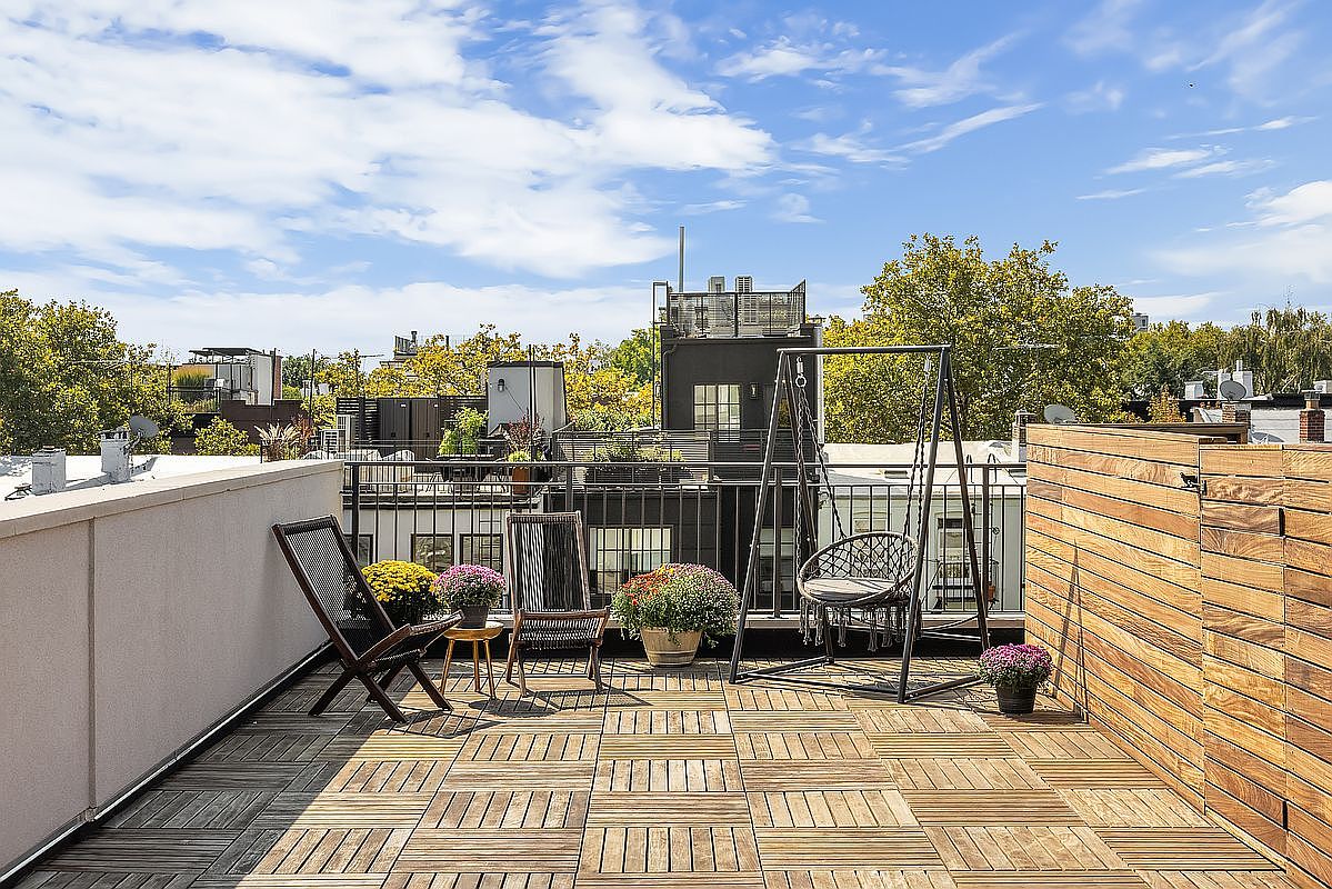 This image showcases a stylish rooftop deck, featuring wooden flooring and a mix of seating options including chairs and a swing. The deck is adorned with potted plants, adding a touch of nature to the urban setting. The view includes surrounding buildings and a bright sky, creating an inviting outdoor living space.
