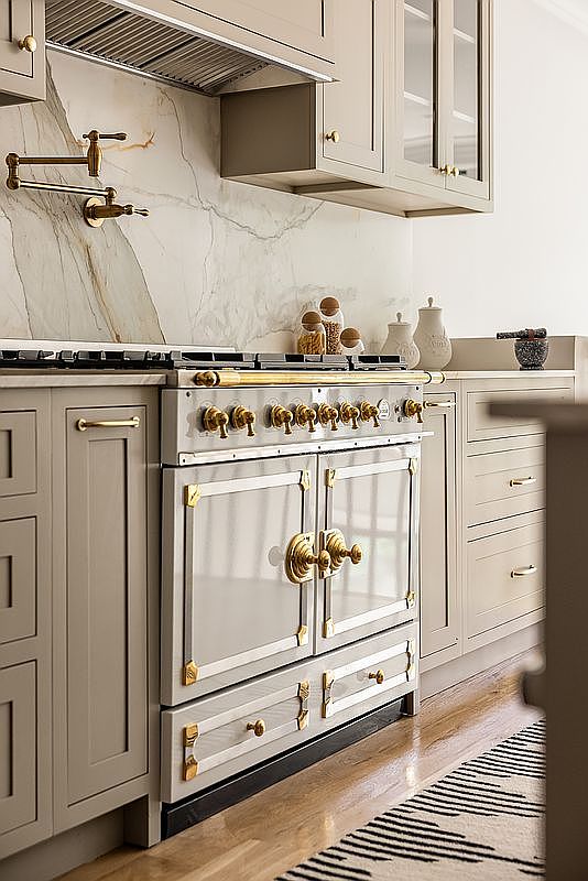 This image showcases a high-end kitchen featuring custom cabinetry in a muted gray tone, accented with elegant gold hardware. A professional-grade range with multiple burners and a decorative backsplash adds a touch of luxury. The kitchen exudes a sophisticated and timeless aesthetic, perfect for discerning buyers.
