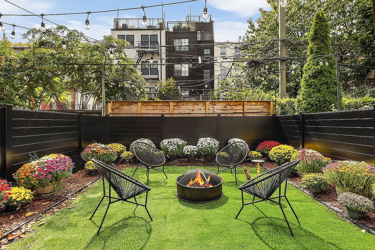 This image showcases a beautifully designed backyard featuring a modern fire pit surrounded by four stylish black chairs, creating an inviting outdoor living space. The yard is adorned with colorful flower arrangements and a well-maintained artificial lawn, enclosed by a sleek, dark-colored fence that provides privacy. String lights overhead add a touch of ambiance, enhancing the appeal for evening gatherings.