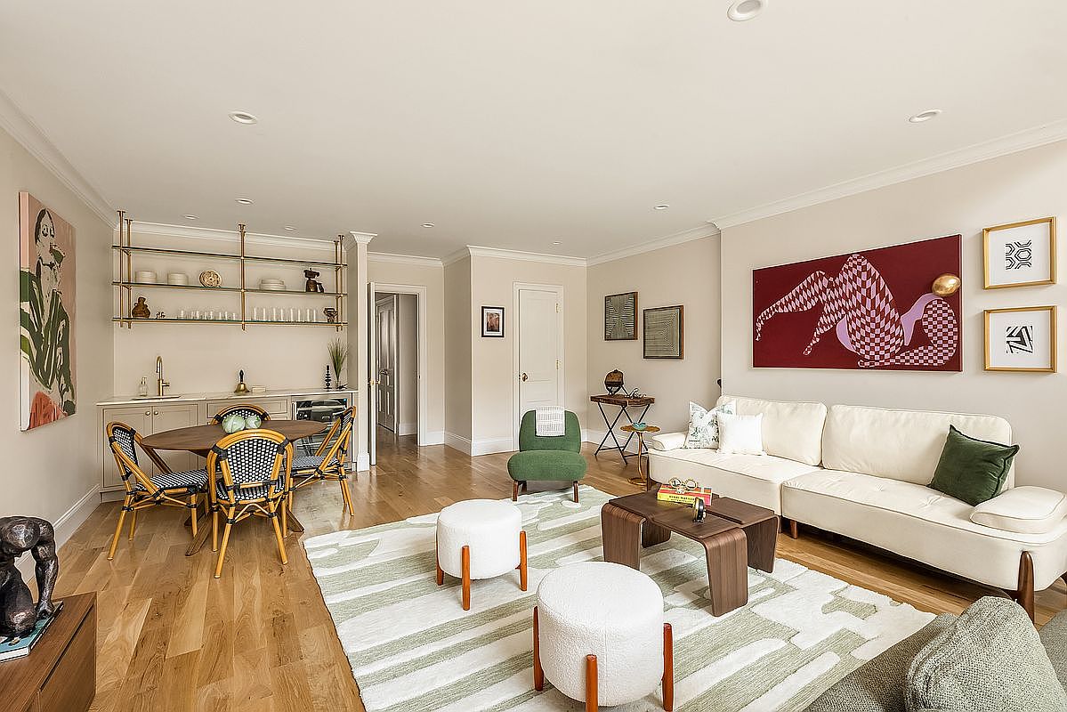 This is an interior shot of a well-decorated living room. The space features a cream-colored sofa, a unique coffee table, and two white ottomans on a patterned rug. Artwork adorns the walls, and a dining area with a built-in bar is visible in the background, creating an inviting and stylish atmosphere.