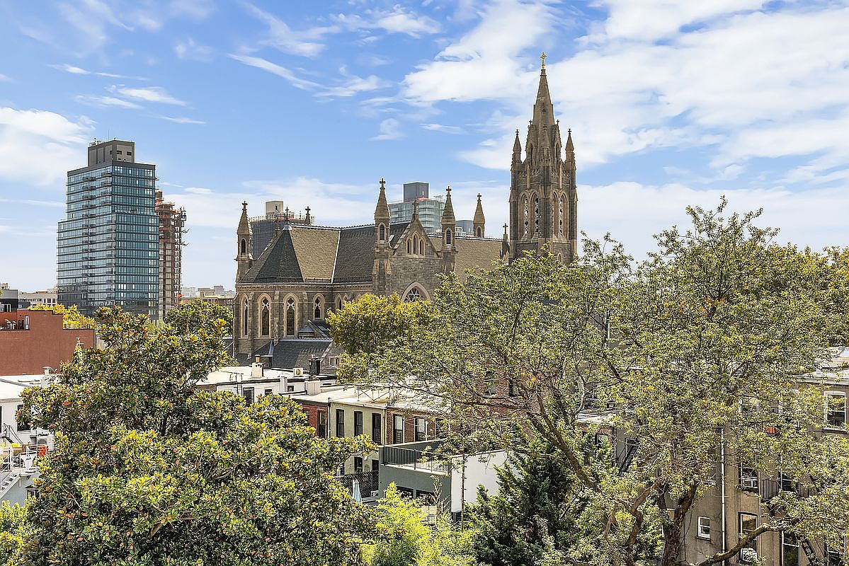 This aerial view showcases a historic church with a tall spire, surrounded by lush green trees and residential buildings. A modern high-rise building is visible in the background, creating a contrast between old and new architecture. The image captures a vibrant urban landscape under a partly cloudy sky.