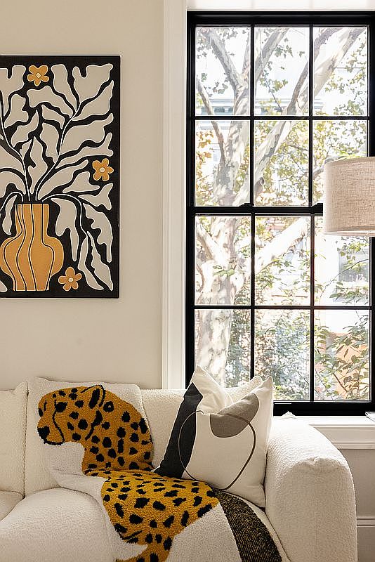 This image showcases a cozy living room corner with a modern aesthetic. A plush, white sofa is adorned with decorative pillows, including a playful leopard-shaped cushion and a geometric design. A striking black-framed window provides natural light and a view of the outdoors, while a contemporary art piece hangs on the wall, adding a touch of sophistication.