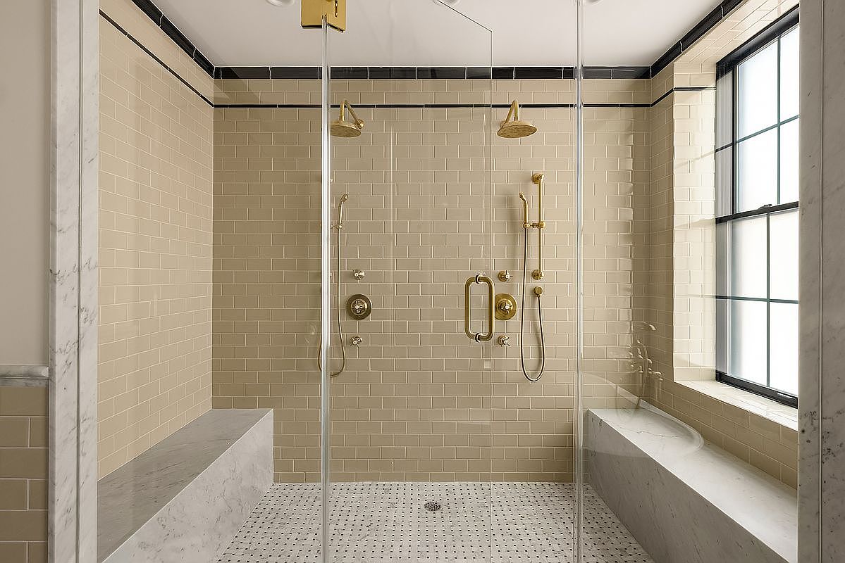 This is a luxurious primary bathroom featuring a spacious walk-in shower with dual shower heads and gold fixtures. The shower has beige subway tile walls, a white and black mosaic tile floor, and marble benches on either side. Glass doors enclose the shower, and a window provides natural light, creating a bright and elegant space.