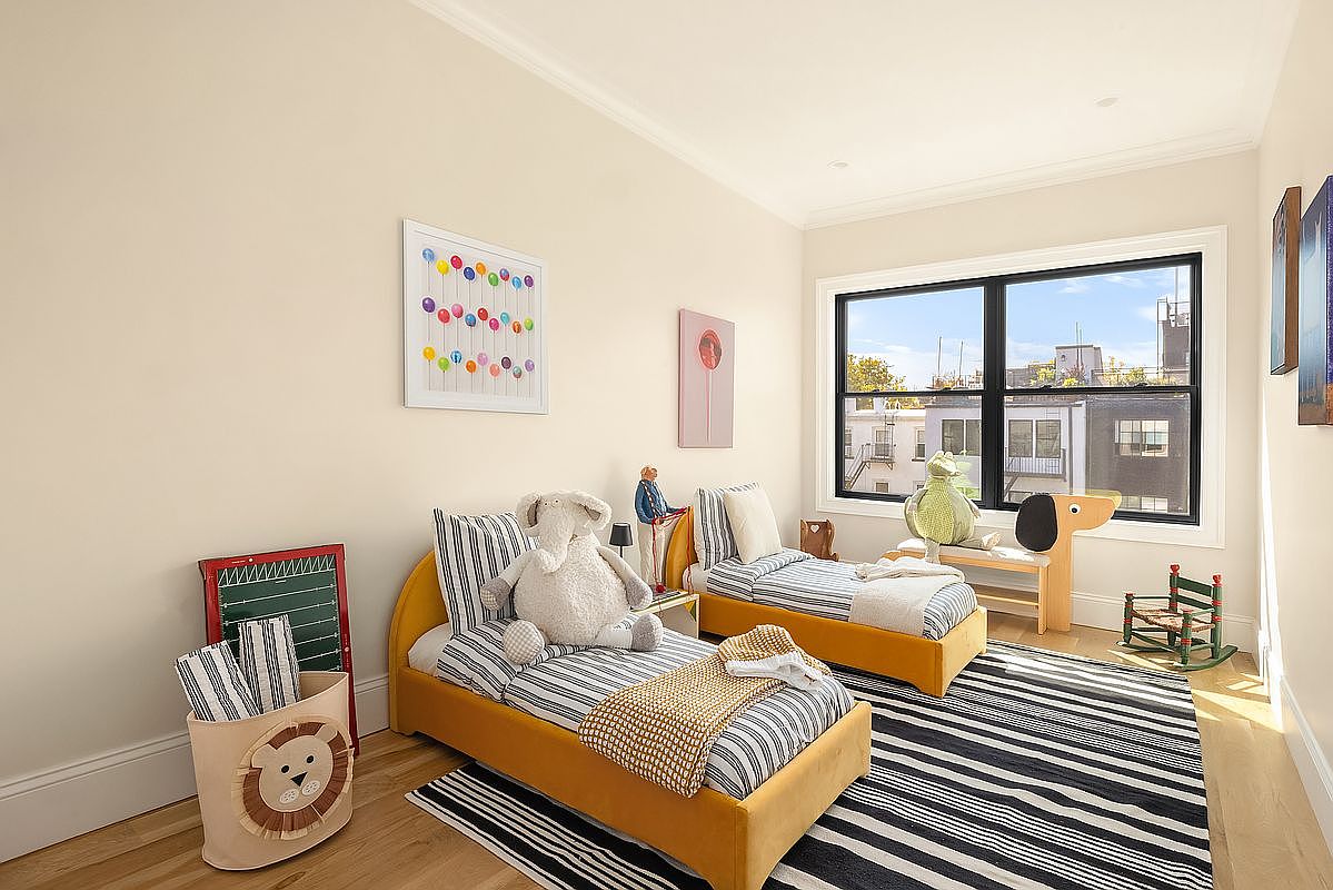This is a charming children's bedroom featuring two twin-sized beds with yellow frames and striped bedding. A black and white striped rug anchors the room, while playful artwork and stuffed animals add a whimsical touch. The room is well-lit by a large window, offering a view of the neighborhood.