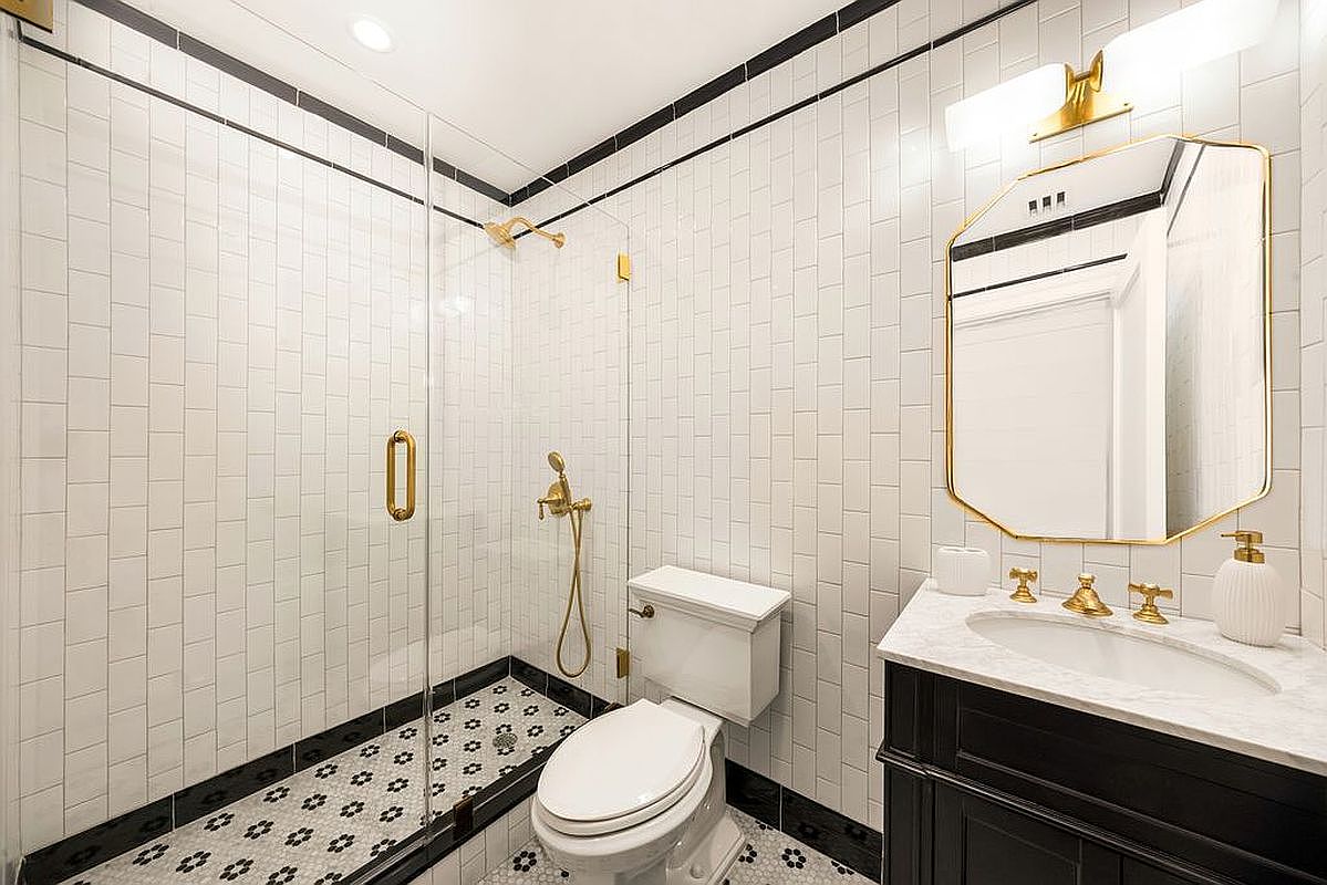 This is a well-lit bathroom featuring white subway tiles with black trim, a black and white patterned tile floor, and gold fixtures. The bathroom includes a glass-enclosed shower, a toilet, and a vanity with a marble countertop and a black cabinet. The mirror has a unique shape with a gold frame, adding a touch of elegance to the space.