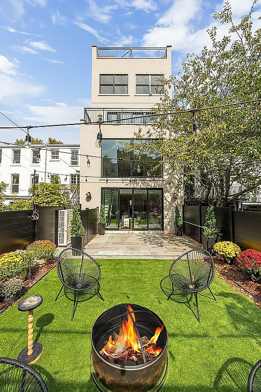 This is a rear view of a modern townhouse backyard featuring a well-manicured lawn, a fire pit, and stylish outdoor seating. The yard is enclosed by a dark fence and adorned with colorful flower beds, creating a cozy and inviting outdoor space. String lights hang overhead, adding a touch of ambiance.
