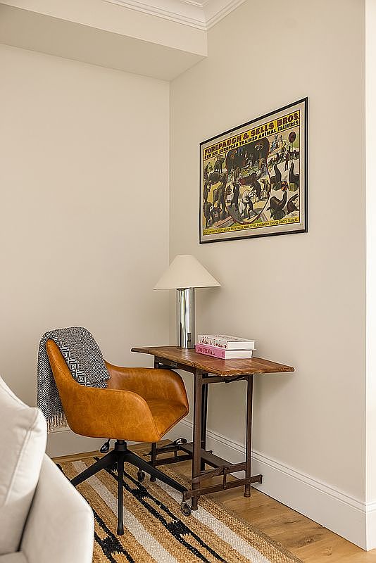 This is an interior shot of a cozy office or study nook. It features a brown leather office chair, a small wooden desk with a lamp and books, and a framed vintage circus poster on the wall. The space is decorated with a neutral color palette and a patterned rug, creating a warm and inviting atmosphere.
