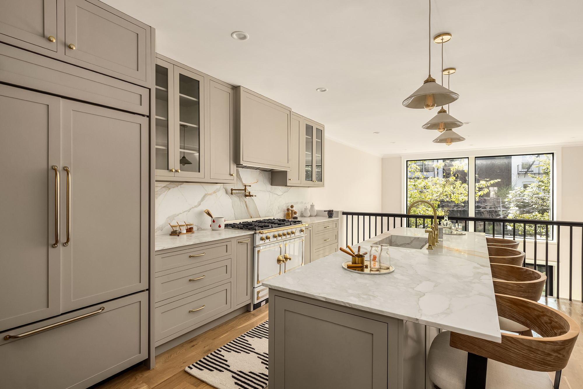 This interior shot showcases a beautifully designed kitchen with gray cabinetry, gold hardware, and marble countertops. The open layout includes a large island with seating, stylish pendant lighting, and high-end appliances, creating an elegant and functional space. A view to a secondary level with a large window adds to the kitchen's appeal.