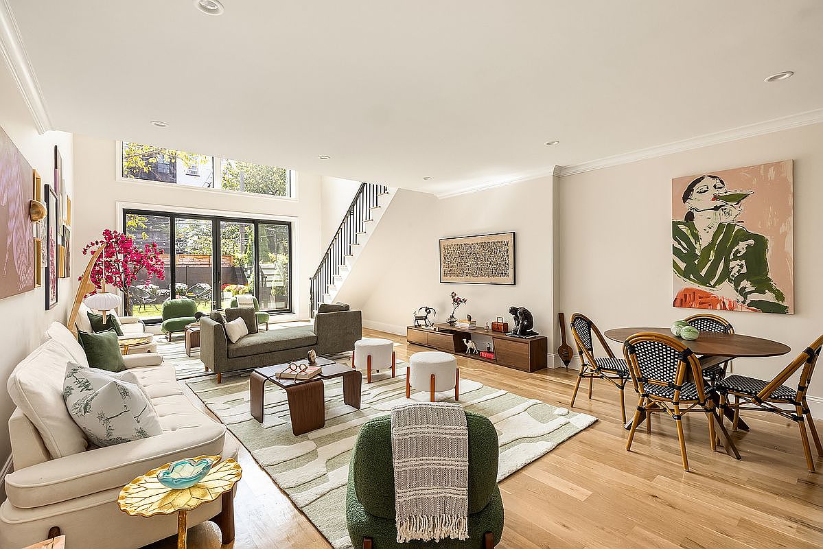 This is a spacious and bright living room featuring a modern design with hardwood floors and neutral-toned walls. The room is furnished with a cream-colored sofa, olive green chaise lounge, and a round dining table with wicker chairs. Large sliding glass doors provide access to the backyard, and artwork adorns the walls, creating a sophisticated and inviting atmosphere.