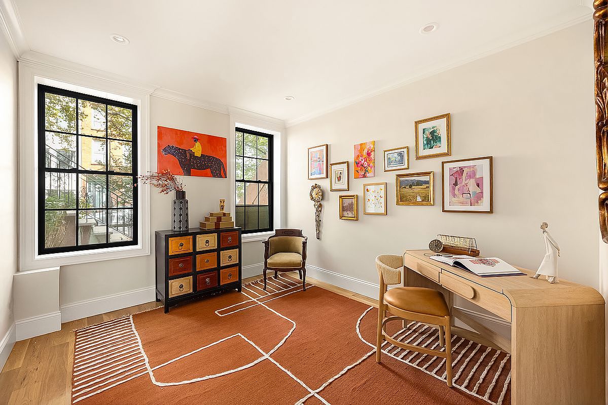 This is an interior shot of a well-decorated office or study. The room features a desk with a chair, a cabinet with drawers, and several framed artworks on the wall. The room is well-lit with natural light coming through the windows, and the overall style is sophisticated and inviting.