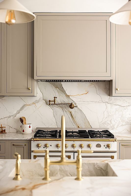 This is a detailed shot of a high-end kitchen featuring custom cabinetry in a neutral tone, complemented by a marble backsplash and countertops with striking veining. A professional-grade range with brass accents and a matching pot filler add a touch of luxury. The focus is on the elegant design and premium materials, creating a sophisticated and inviting culinary space.
