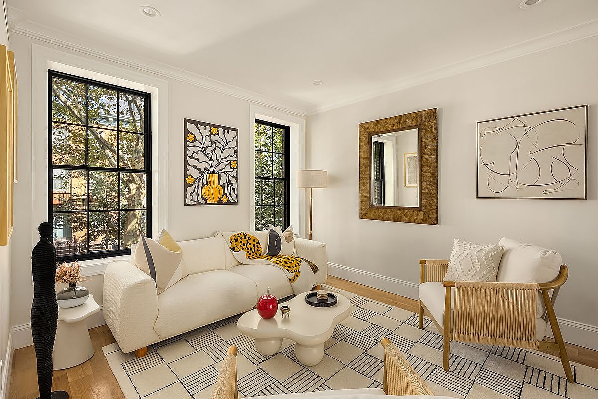This is a bright and stylish living room featuring a cream-colored sofa, two woven armchairs, and a unique white coffee table. The room is decorated with modern art, including framed prints and a decorative mirror, and a patterned rug adds texture to the space. Large windows provide natural light and views of the outdoors, creating a welcoming and sophisticated atmosphere.