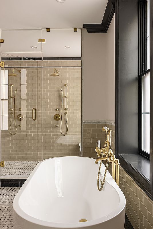 This is a luxurious primary bathroom featuring a freestanding white bathtub with gold fixtures, complemented by a glass-enclosed shower with dual showerheads and matching gold hardware. The walls are tiled in a neutral tone, and a dark-framed window provides natural light. The overall impression is elegant and modern.