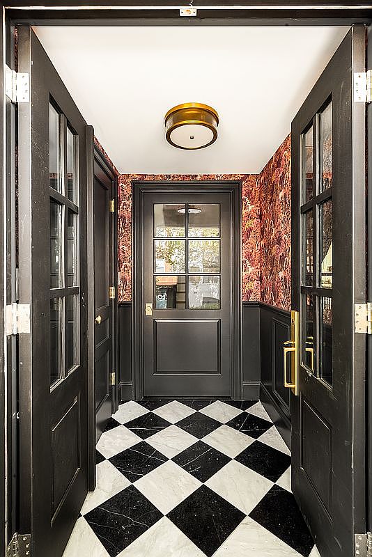 This interior shot showcases a stylish entryway with black and white checkered marble flooring. The walls are adorned with patterned wallpaper above black wainscoting, and a black door with glass panes leads further into the home. Two black doors with glass panes are open on either side of the hallway, creating a welcoming and sophisticated atmosphere.