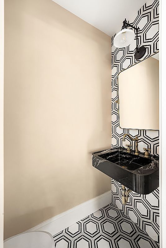 This is a stylish guest bathroom featuring a black marble sink with gold fixtures, set against a backdrop of geometric black and white wallpaper and matching floor tiles. A vintage-style light fixture and a frameless mirror add to the room's sophisticated and modern aesthetic. The walls are painted a neutral beige color.