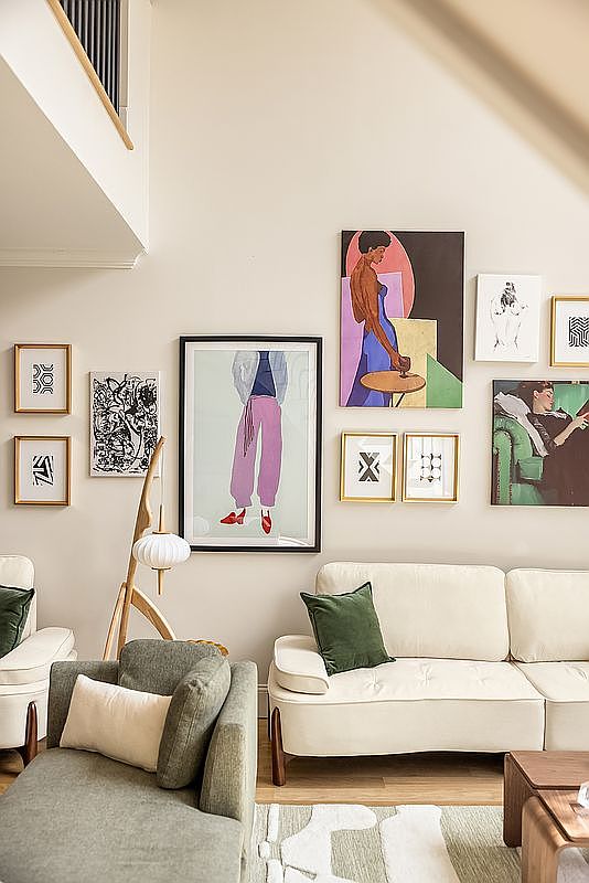 This is an interior shot of a stylish living room featuring a gallery wall with various framed artworks. The room is furnished with a cream-colored sofa, a green armchair, and a modern floor lamp, creating a cozy and artistic atmosphere. The neutral color palette and the arrangement of furniture suggest a comfortable and inviting space, perfect for relaxation and entertainment.