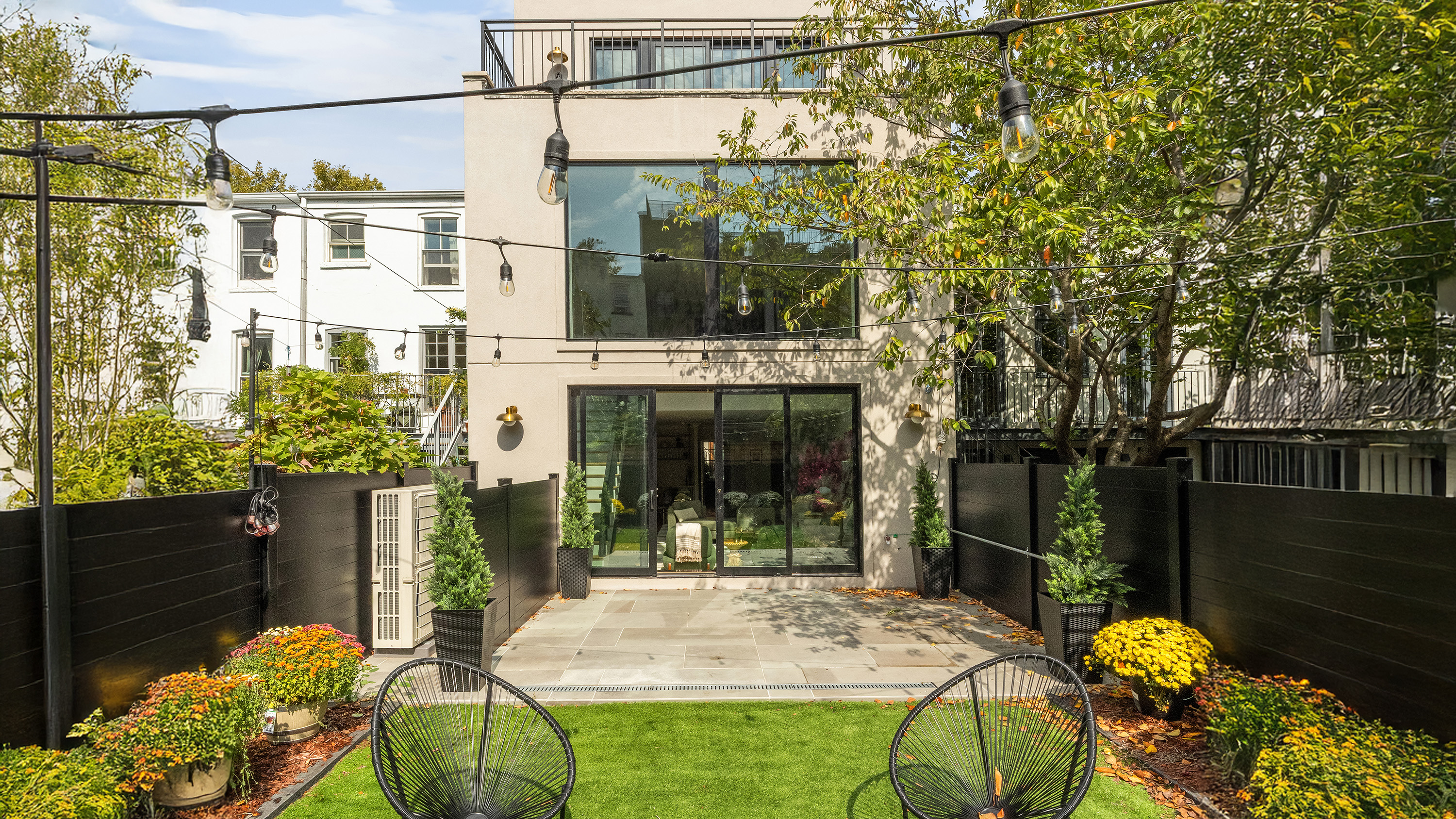 This is a rear view of a beautifully landscaped yard, perfect for outdoor living. The garden features a manicured lawn, patio area, and elegant black fencing, enhancing privacy and setting a sophisticated tone. String lights are hung above and add to the ambiance for evening gatherings.