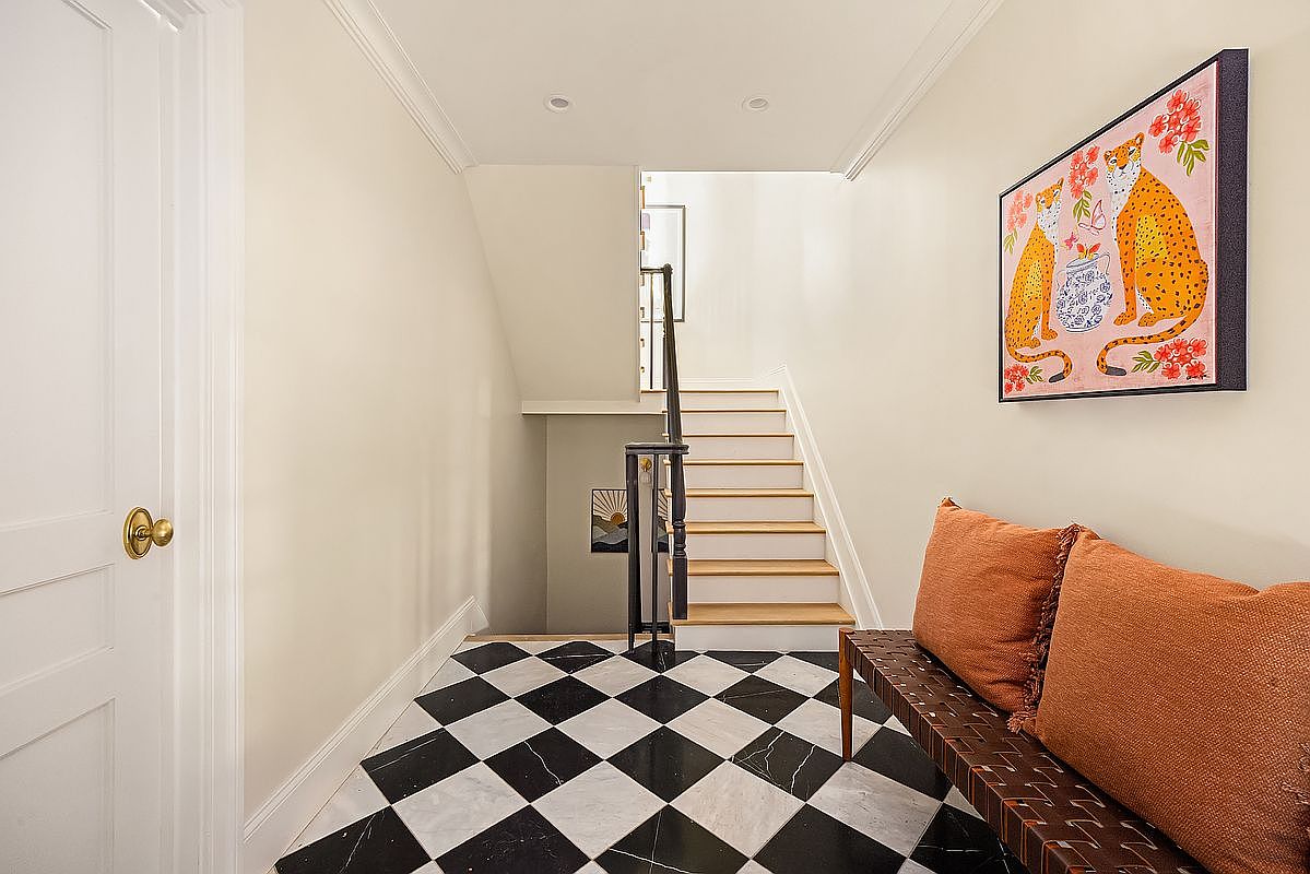 This interior shot showcases a stylish hallway with black and white checkered marble flooring leading to a staircase. A woven bench with orange pillows sits against the wall, complemented by a framed artwork featuring leopards and flowers. The walls are painted in a neutral tone, and a white door is visible on the left, creating a bright and inviting entryway.