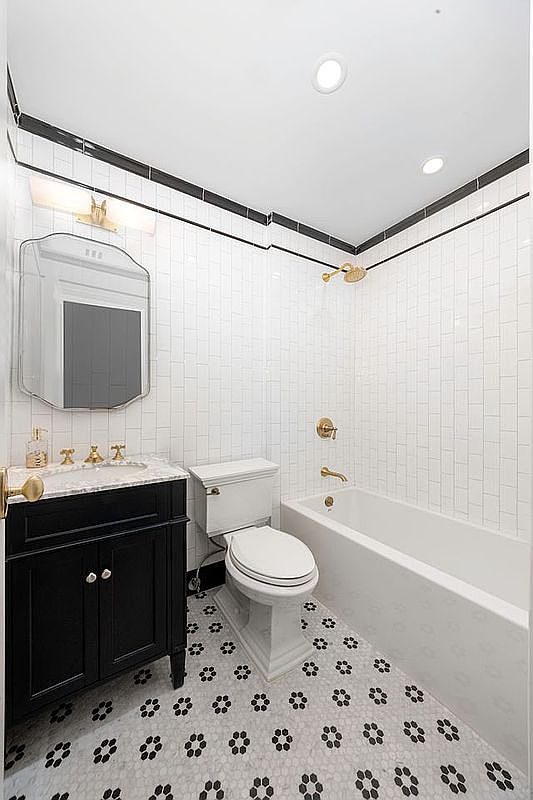 This is a well-lit bathroom featuring a black vanity with a marble countertop, a classic white toilet, and a bathtub with white subway tile surround and gold fixtures. The floor is decorated with a black and white floral pattern. The bathroom has a clean and traditional style.
