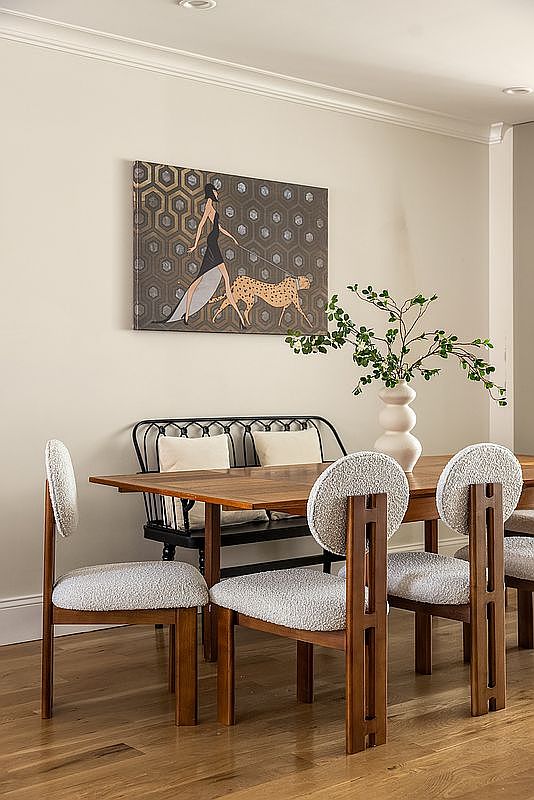 This interior shot showcases a dining room with a wooden table and unique chairs featuring rounded, upholstered backs. A black bench with cushions sits against the wall beneath a modern art piece depicting a woman and a cheetah. A vase with greenery adds a touch of nature to the space, creating a stylish and inviting atmosphere.