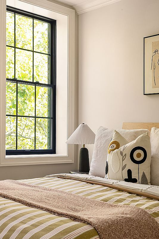 This is a cozy bedroom featuring a bed with striped bedding and decorative pillows. A black-framed window provides natural light, and a bedside lamp adds a warm glow. The room has a serene and inviting atmosphere, perfect for relaxation.