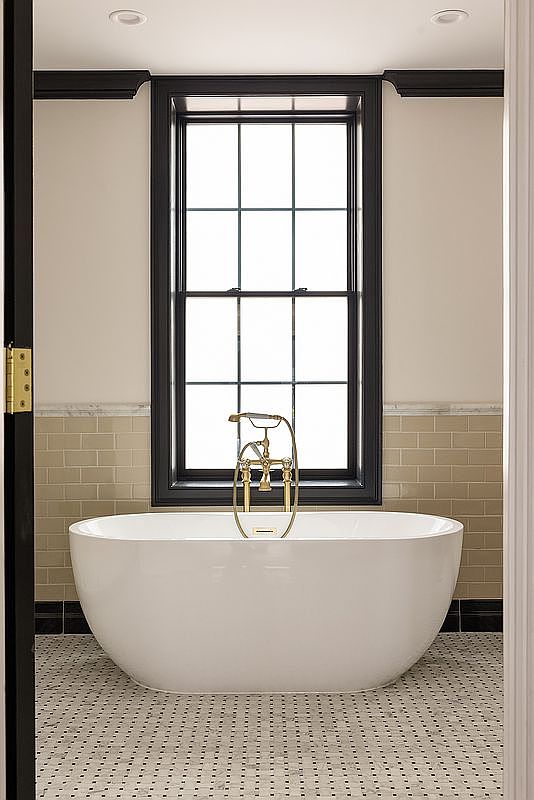 This is a well-lit primary bathroom featuring a freestanding white bathtub with gold fixtures positioned in front of a window with black trim. The walls are a neutral beige color with a tile accent, and the floor is covered in a small, patterned tile. The overall impression is clean, modern, and luxurious.