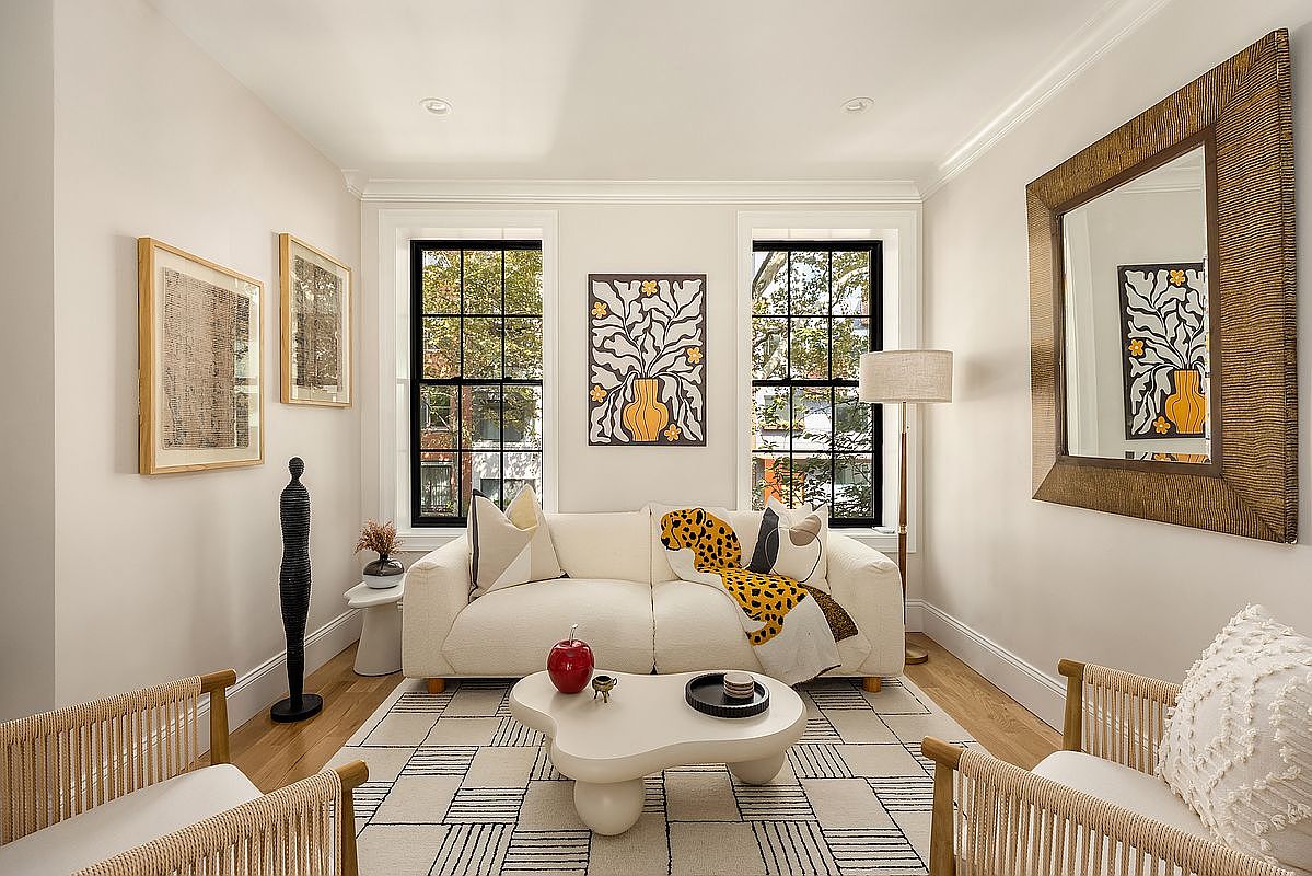 This is a bright and stylish living room featuring a neutral color palette with pops of color from artwork and accessories. The room is well-lit with natural light from two windows and features a comfortable white sofa, a unique coffee table, and decorative art pieces. The overall impression is modern, inviting, and well-designed, perfect for relaxing or entertaining.