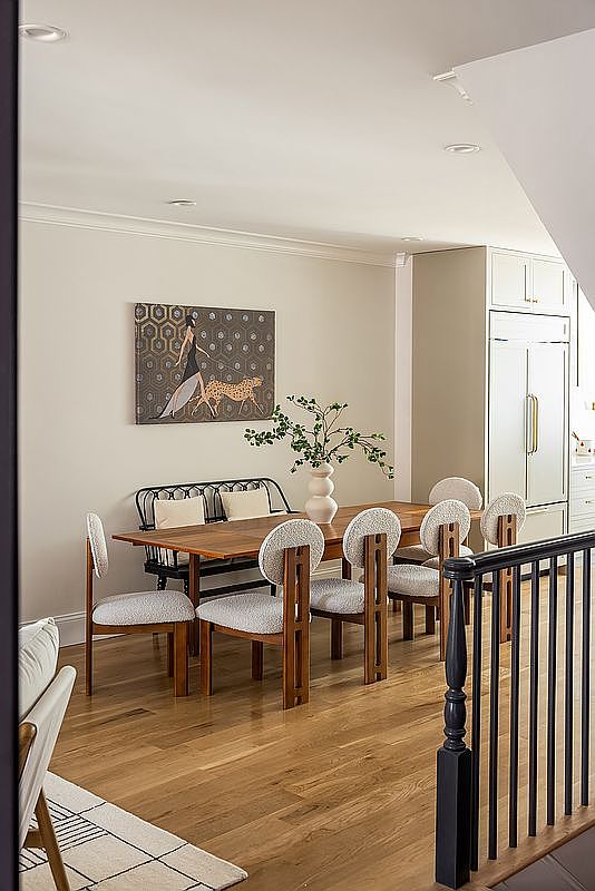 This is an interior shot of a dining room featuring a wooden table with six chairs and a bench. A modern art piece hangs on the wall above the bench, and a vase with greenery sits on the table. The room has hardwood floors and neutral-colored walls, creating a warm and inviting atmosphere.