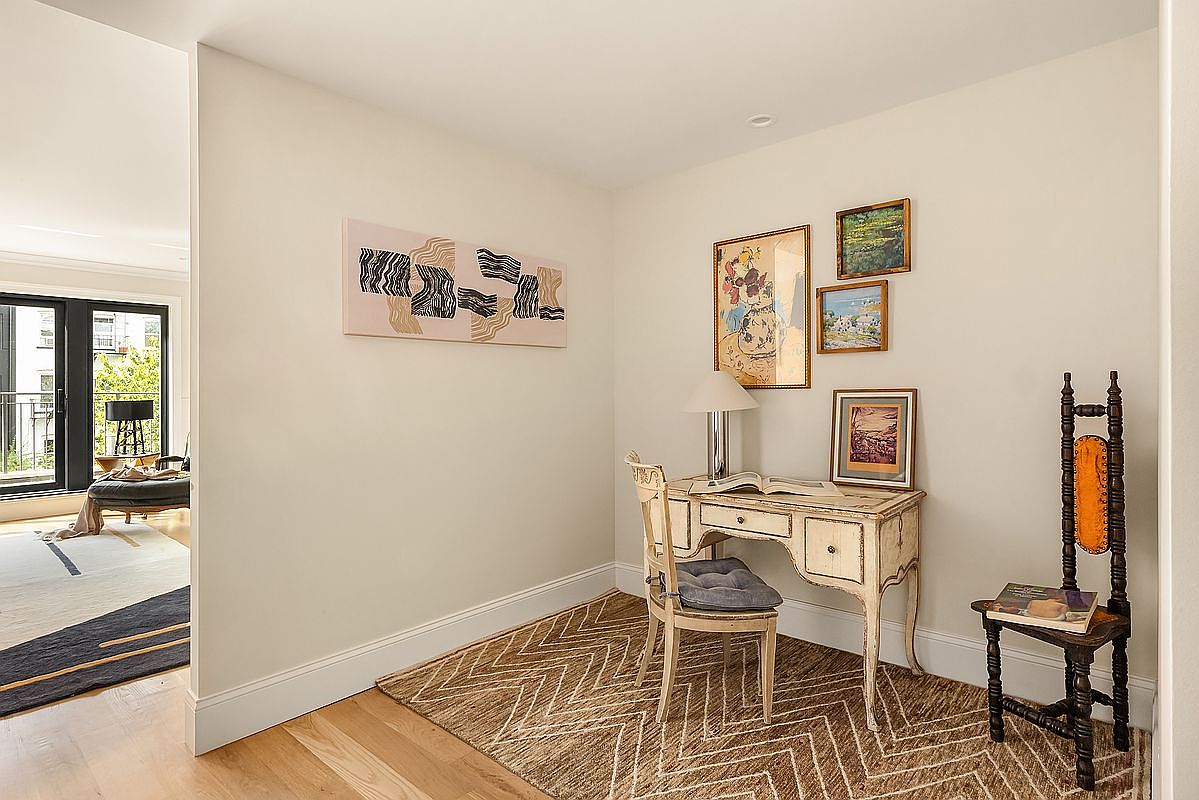 This interior shot showcases a cozy office or study area featuring a vintage-style desk and chair, complemented by artwork and a decorative rug. The room is bathed in soft, natural light, creating a warm and inviting atmosphere. A glimpse into an adjacent living area with a balcony adds depth and suggests a seamless flow within the home.