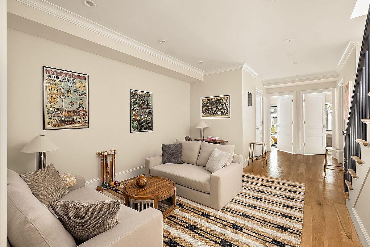 This is an inviting living room featuring neutral-toned sofas, a round wooden coffee table, and a striped rug that adds texture to the space. Artwork adorns the walls, and the hardwood flooring extends into the hallway, creating a seamless transition. The room is well-lit and appears cozy and comfortable, perfect for relaxation and entertaining.