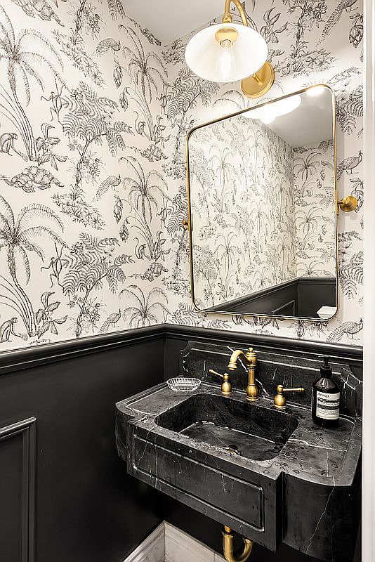 This is a stylish guest bathroom featuring a black marble sink with gold fixtures, complemented by black wainscoting and a whimsical black and white wallpaper with a tropical motif. A gold-framed mirror and sconce add to the luxurious feel of the space. The perspective is a medium shot, highlighting the sink area and the unique design elements.