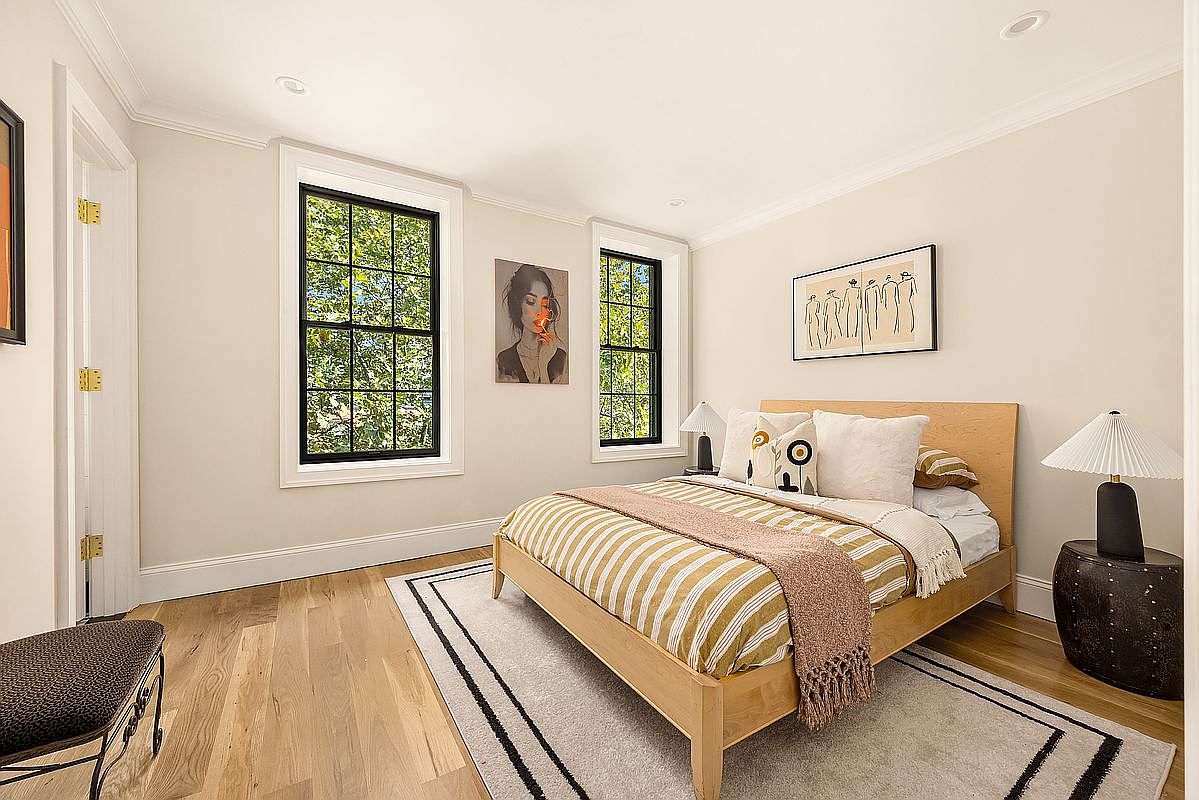This is a bright and inviting guest bedroom featuring hardwood floors, two windows with black frames offering natural light, and a comfortable bed with striped bedding and a decorative throw. Artwork adorns the walls, complementing the neutral color scheme and creating a serene atmosphere. A small side table with a lamp adds a touch of elegance.