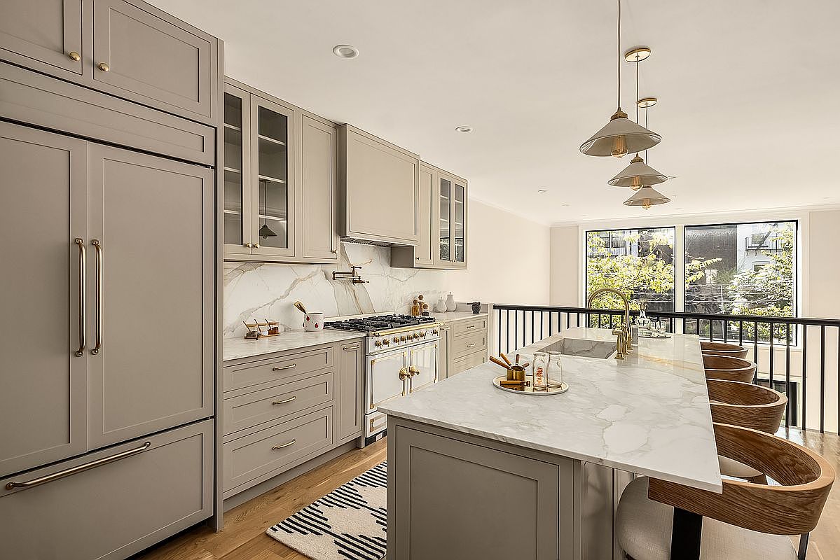 This is a well-lit kitchen featuring gray cabinetry, marble countertops, and stainless steel appliances. A kitchen island with bar seating is present, illuminated by pendant lights. The overall design is modern and elegant, creating a welcoming and functional space.
