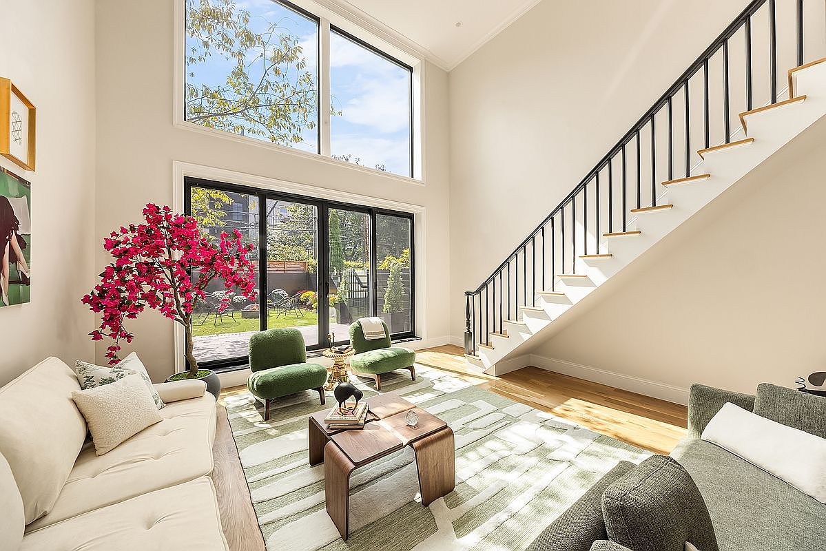 This is a bright and airy living room featuring a modern staircase, large windows overlooking a garden, and contemporary furniture. The room is decorated in neutral tones with pops of green and pink from the plants and chairs, creating a welcoming and stylish atmosphere. The hardwood floors and area rug add warmth and texture to the space.