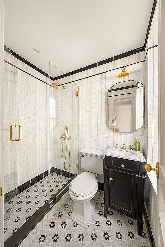 This is a well-lit bathroom featuring a glass-enclosed shower with gold fixtures, a white toilet, and a dark vanity with a marble countertop. The walls are tiled in white with a black accent stripe, and the floor is covered in a patterned tile. The overall impression is clean, modern, and stylish.