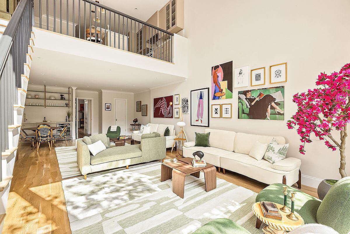 This is a bright and airy living room featuring a double-height ceiling and a gallery wall with various art pieces. The room is furnished with a light green sofa, a cream-colored sofa, and a unique wooden coffee table, all set on a patterned rug. A staircase with a black railing leads to a loft area, and a large pink bougainvillea adds a pop of color to the space.