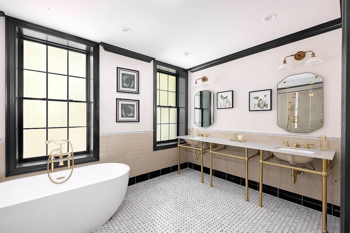 This is a well-lit primary bathroom featuring a freestanding white bathtub with gold fixtures, a double vanity with marble countertops and gold legs, and black-framed windows. The walls are painted a light neutral color, complemented by a beige tile wainscoting and black trim, while the floor is covered in a classic black and white mosaic tile pattern. The perspective is from the doorway, showcasing the entire space.