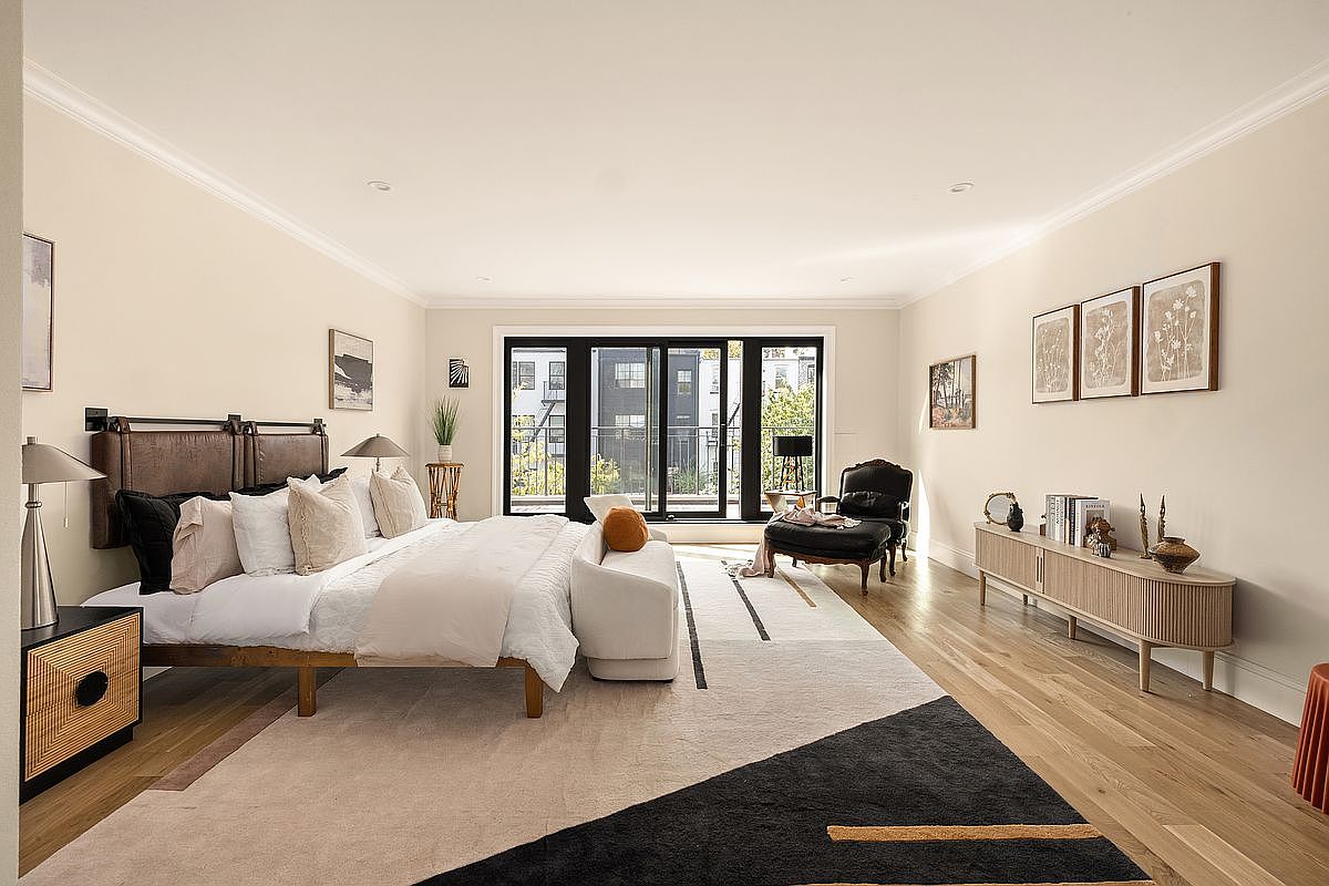 This is a spacious primary bedroom featuring a large bed with a leather headboard and neutral-toned bedding. The room is well-lit with natural light streaming in from a large sliding glass door that leads to a balcony. The decor includes modern furniture, artwork, and a stylish area rug, creating a comfortable and inviting atmosphere.