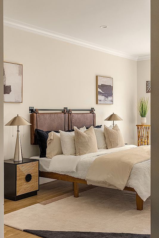 This is a well-lit primary bedroom featuring a bed with a brown leather headboard and neutral-toned pillows and bedding. A modern nightstand with a lamp sits beside the bed, and artwork adorns the walls. The room has a calm and inviting atmosphere, perfect for relaxation.